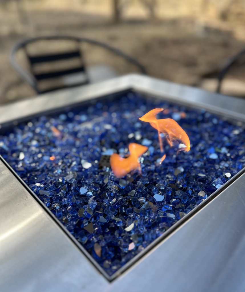 A close-up of a fire pit filled with blue glass rocks. Orange flames rise from the center. Metal edges frame the square pit, and blurred chairs are visible in the background.